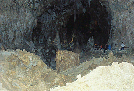 [There is an entrance into a dark hole in the stone. The railing and people on the right are quite tiny in comparison to the size of the hole. There is a multitude of rock crevices and pieces in the area that is not the walkway.]